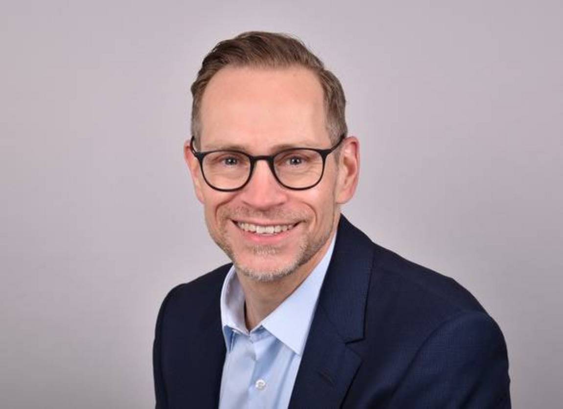 Lächelnder Mann mit kurzen hellbraunen Haaren, Brille und gestutztem Bart, der eine marineblaue Anzugsjacke und ein hellblaues Hemd trägt und vor einem schlichten hellgrauen Hintergrund posiert.